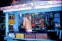 Another waits for customers at right. Note the two young Moplah boys going by dressed in their traditional attire.